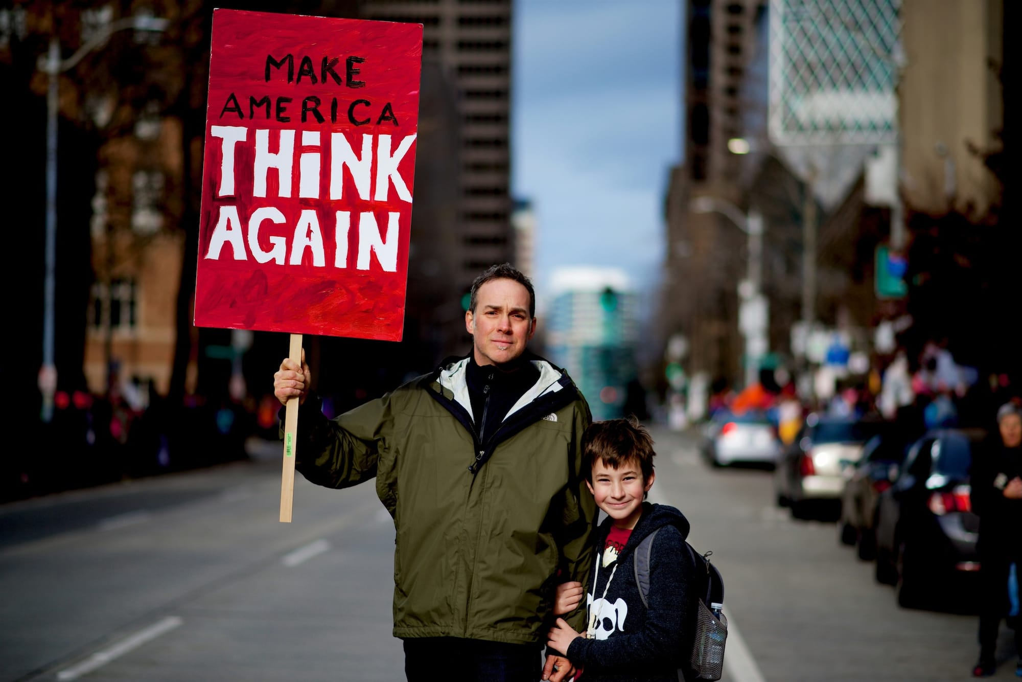 A man and his son on a US street holding a placard reading Make America Think Again